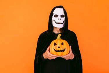 Person with skeleton mask, cape and hood, holding a pumpkin jack-o'-lantern, dressed up for Halloween on orange background