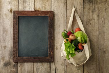 Fresh salad vegetables in a natural cotton reusable bag hanging next to a wooden framed blank blackboard on an old wooden plank wall.
