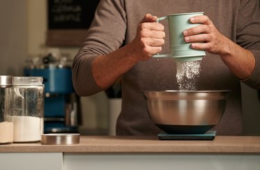 man pouring flour through a flour sifter into a metal bowl on a digital scale
