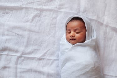 a one month old baby sleeping and swaddled in white cloth lying in white cloth background.