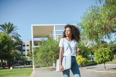 Portrait of young woman with mobile phone and laptop bag walking down the street looking at cell phone