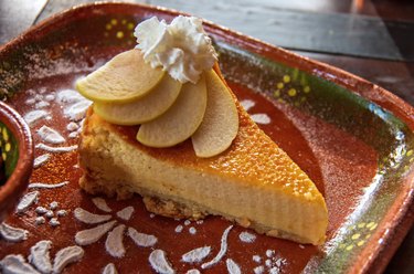 Slice of baked cheesecake, garnished with sliced apple and cream on a traditional clay plate with a stenciled icing sugar floral design