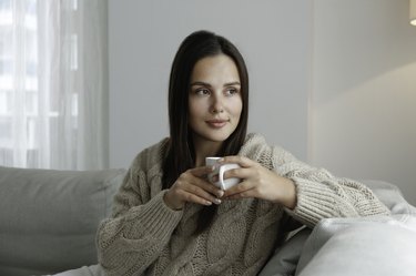 Young woman sitting on a gray sofa  wearing a knit sweater with a porcelain coffee cup in hand.