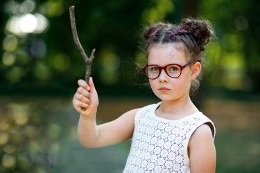 Funny adorable little kid girl with glasses and wooden magic wand playing Harry Potter in park on sunny day.