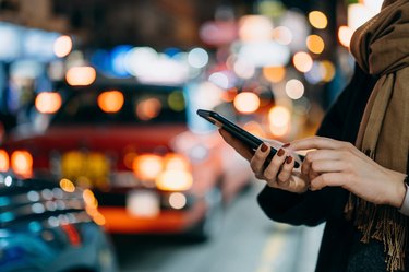 Close up of young woman using mobile app device on smartphone to arrange taxi ride in downtown city street, with illuminated busy city traffic scene during rush hour with traffic congestion in the evening
