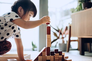 Creative little Asian girl crouching on the floor playing with wooden building blocks at home