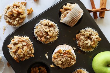 Image of muffin baking tray with rows of homemade apple muffins in paper cake cases topped with streusel crumble on marble effect background, elevated view