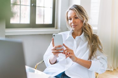 Young smiling blonde woman with long hair in white shirt using smartphone in the bright modern office, woman send message by smartphone