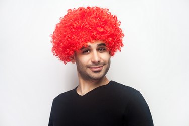 Portrait of funny stylish hipster man with curly red wig smiling at camera, looking playful positive. white background