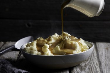 Container of gravy being poured onto bowl of mashed potatoes