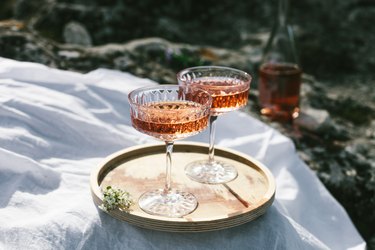 Two glasses of rose wine outdoors on wooden tray and white textile, picnic in spring. Sunny day, green grass and blossoming cherry trees.