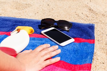 Cropped Hand By Mobile Phone On Towel At Beach During Sunny Day