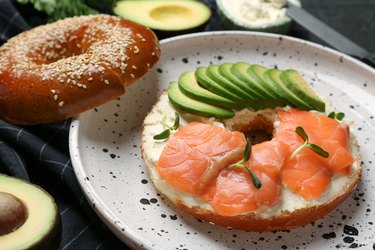 Delicious bagel with cream cheese, salmon and avocado on plate, closeup
