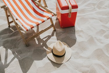 Overhead view of a relaxing beach scene, including a deck chair, sun hat and cool box.