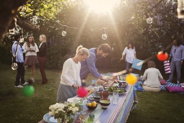 Man and woman preparing food at dining table with friends in background during summer party