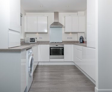 A Modern new build kitchen with built in units and Oven with Aluminium Cooker Hood."nFinished in high gloss White with under cabinet lights and grey wooden flooring.