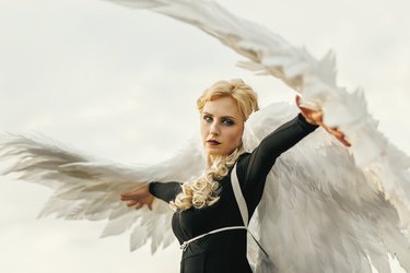 Young glamorous woman in a black dress with a large white angel wings on the background of a dramatic sunset sky. The concept of a fallen dark angel