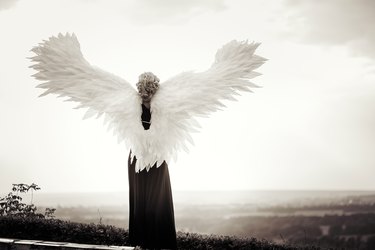 Young glamorous woman in a black dress with a large white angel wings on the background of a dramatic sunset sky. The concept of a fallen dark angel
