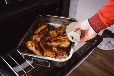 Male hand removing a shallow stainless steel roaster, filled with roasted chicken legs, from a domestic oven
