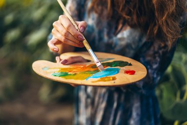 Close up hands of female artist holding brush and palette with oil paints. Blurred background with easel in sunflower field