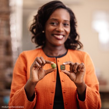 Chef Selassie Atadika holds two chocolate truffles and wears a bright orange jacket