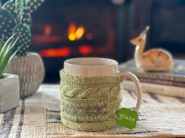 Light green mug cozy with tea bag in front of a fireplace