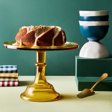 Yellow clear glass cake stand with a bundt cake on top of it against a teal green background.