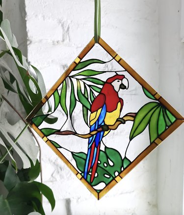 Sun catcher kit for adults from Etsy. It's a large, diamond-shaped painting panel of a colorful parrot surrounded by greenery with plenty of negative space for light to come through.