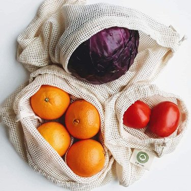 Three mesh cotton reusable produce bags containing oranges, tomatoes, and purple cabbage.