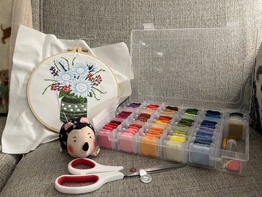 Embroidery supplies laid on a coach, including a clear plastic container of thread, fabric scissors, a silver needle threader, a hedgehog pincushion and a partially finished embroidery piece
