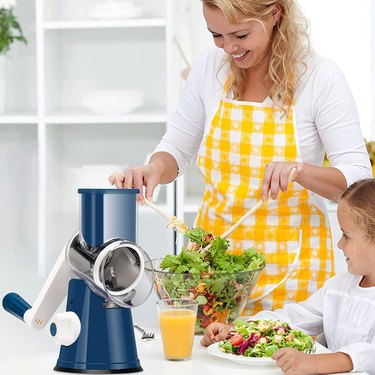 Smiling woman in yellow apron tossing salad next to blue Ourokhome mandoline slicer.