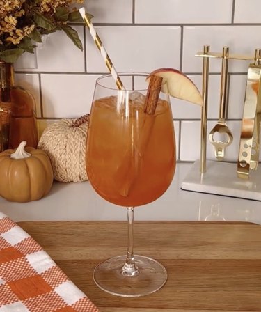 Glass filled with reddish orange cocktail, a cinnamon stick, striped straw and apple slice