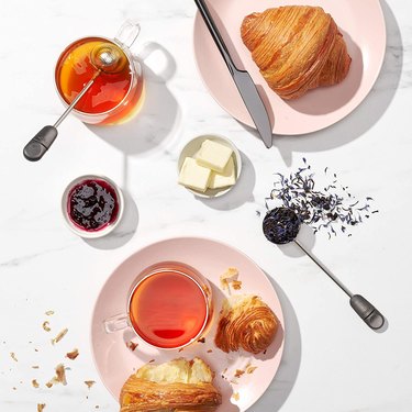 OXO BREW Twisting Tea Ball Infuser on a marble countertop surrounded by loose leaf tea and croissants.