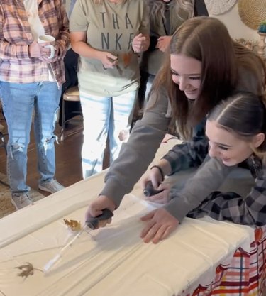 Two women use turkey basters to blow leaves down a tabletop