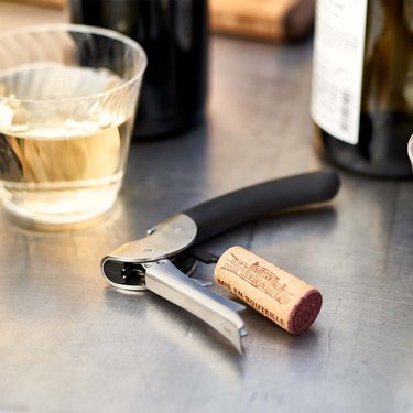 OXO wine bottle opener on a steel countertop with a cork and glass of wine.
