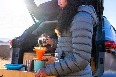 Woman in parka using orange pour-over coffee maker out of the back of an SUV.
