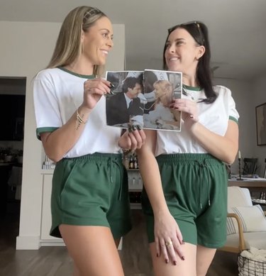 Two women dressed in forest green shorts and white and green T-shirts holding a torn-in-half photo of a bride and groom