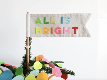 All is Bright multicolor flag tree topper on a wooden dowel.