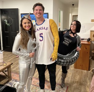 Woman in black shirt reading "Official Third Wheel" and inflatable wheel costume stands behind a couple posing for Halloween