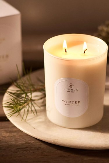 A two-wick candle with a label that says 'Winter' on a round plate with a sprig of pine.