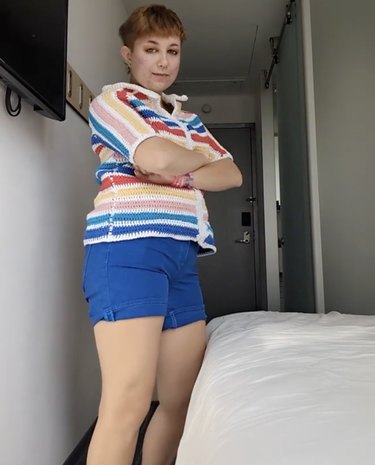 Person with short red hair dressed in a multicolored short-sleeve sweater top and blue shorts