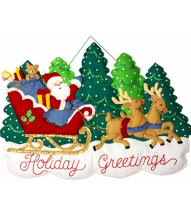 A plush wall hanging featuring Santa in his sleigh with green Christmas trees behind him and the words "Holiday Greetings" in script across the bottom.