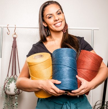 Macrame artist Casey Alberti in her studio holding three giant spools of thread in yellow, blue and red