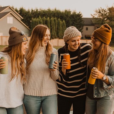 Group of 4 women in sweaters and knit hats each holding a BrüMate Hopsulator Slim Insulated Can Cooler in different colors.