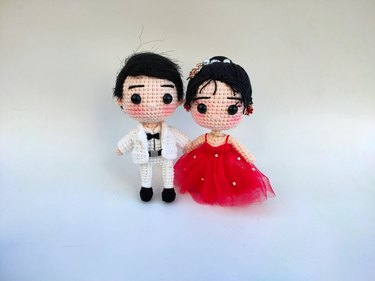 A set of crochet dolls. The doll on the left has short black hair and wears a white suit, while the doll on the right has longer black hair and wears a red dress with white beads.
