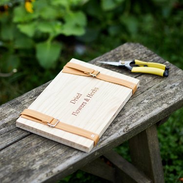A wooden flower press with light brown leather straps sitting on a rustic bench next to flower clippers