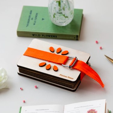 A small rectangle flower press with a bright orange vertical strap, sitting next to a green wildflowers book