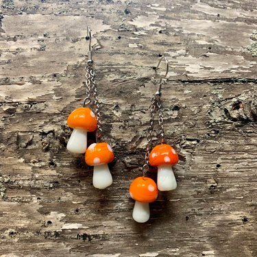 A set of earrings with two orange mushrooms dangling from each earring. The mushrooms feature white dots.