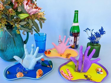 Three trays sit near a green wine bottle and flower vases, each tray with a realistic 3D-printed human hand. The trays are yellow and purple, pink and light orange and blue.