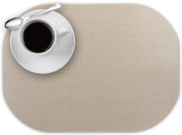 Andie Home oval placemat with a fabric-look print, shown in an overhead view with a cup of coffee and saucer in the top left corner